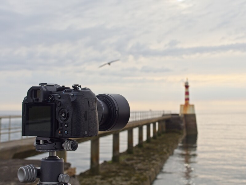 Camera on tripod aimed at a pier extending into calm water with a striped lighthouse beacon in the distance.