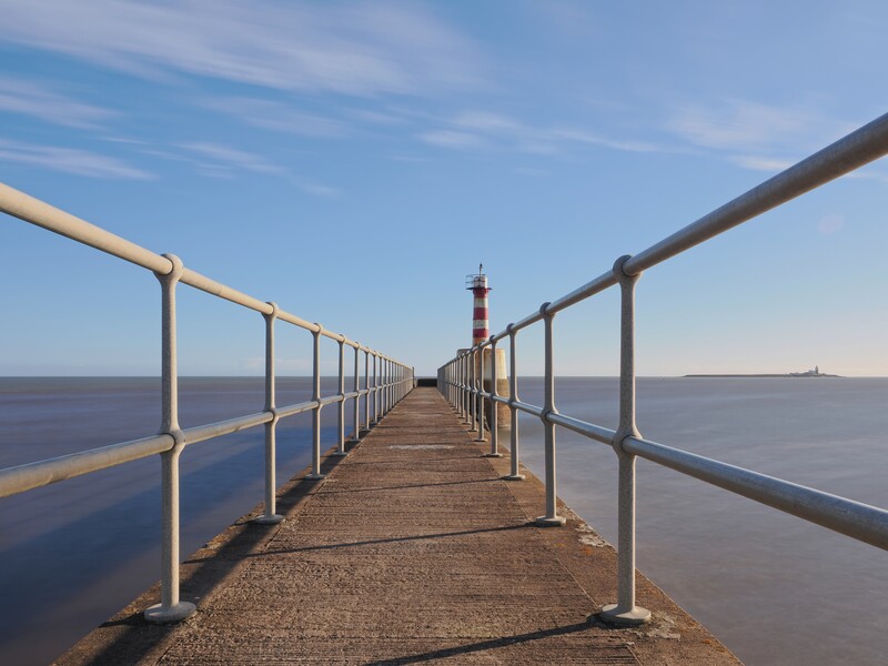 Long wooden pier with white railings extending toward a distant lighthouse under clear blue sky.