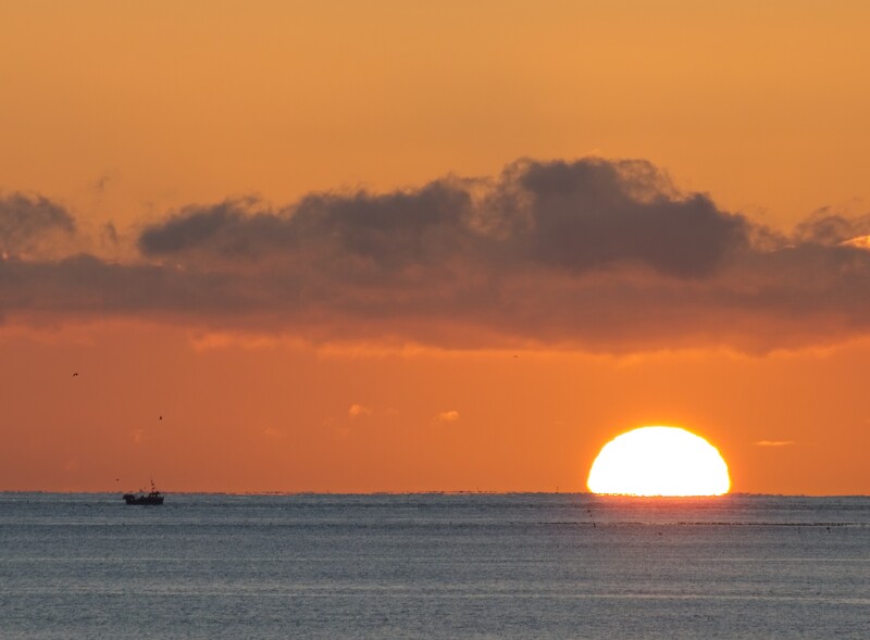 Large sun touching the horizon at sunset over calm ocean water with a small fishing boat silhouetted on the left.