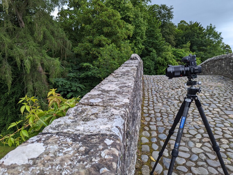 Professional camera on tripod positioned on a cobblestone surface beside a weathered stone wall overlooking green landscape.