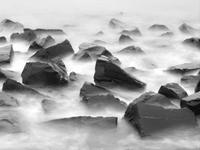 Rocks partially submerged in flowing water with soft, misty appearance from long exposure.