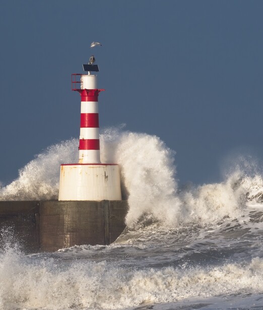 Red and white striped lighthouse surrounded by massive crashing waves during storm conditions.