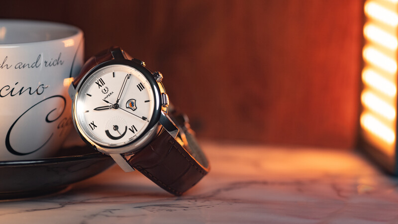 Luxury chronograph watch with black leather strap displayed next to a ceramic mug on marble surface under warm golden lighting.