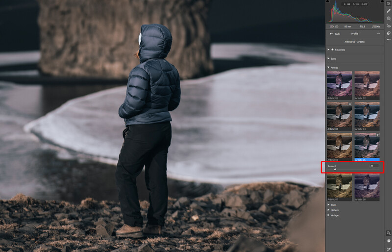 Person in dark puffy jacket standing on rocky terrain overlooking a misty lake and forested landscape.