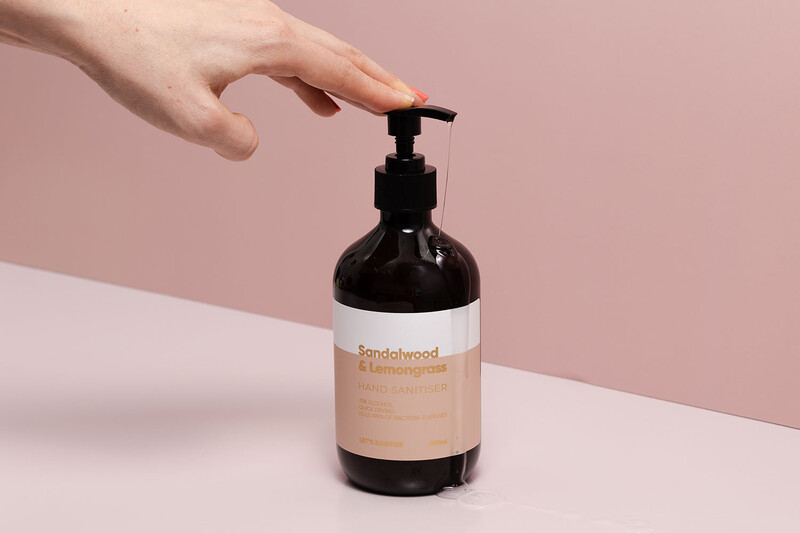 Hand reaching toward a dark glass pump bottle with a tan label against a pink and white background.