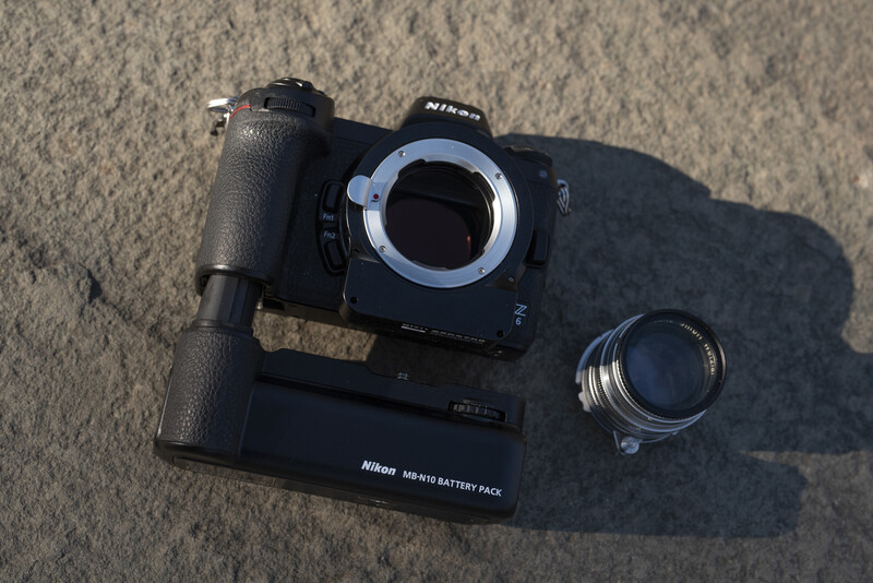 Nikon camera body with battery grip, lens, and accessories laid out on concrete.