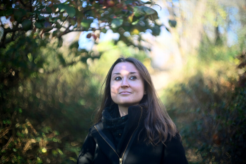 Woman with long dark hair smiling at camera among green foliage in natural daylight.