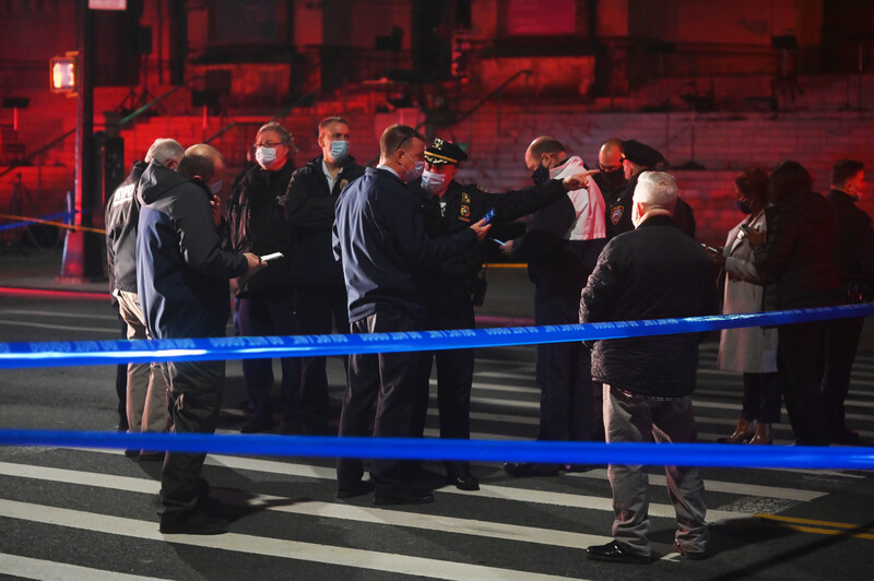 Group of officials and media gathered behind blue police tape at a nighttime street scene with red lighting.