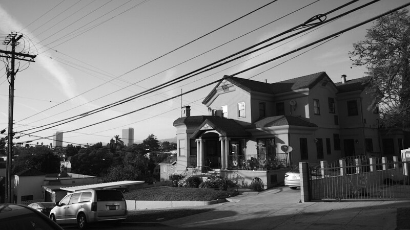 Black and white photograph of a two-story Victorian house with a front porch and white railings, parked car visible on the street.