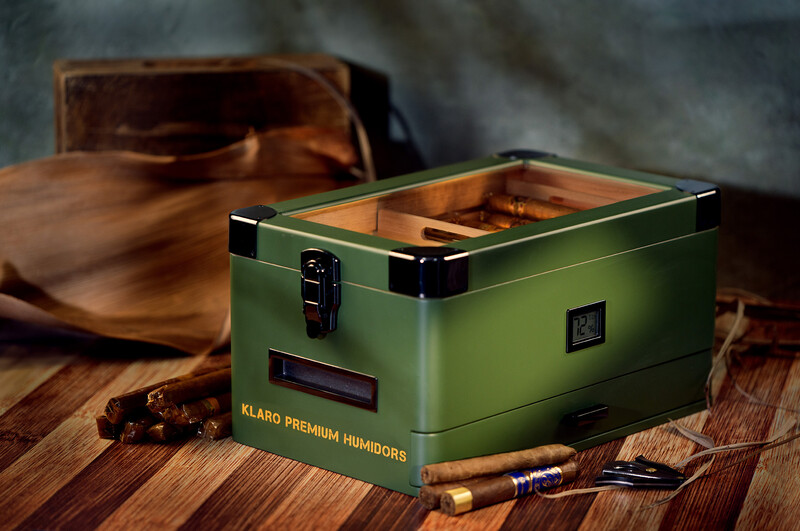 Green metal humidor with open lid displaying interior compartment and cigars beside it on wooden surface.