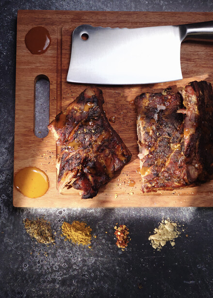 Two cooked steaks on a wooden cutting board with knife, white plate, and spice bowls.