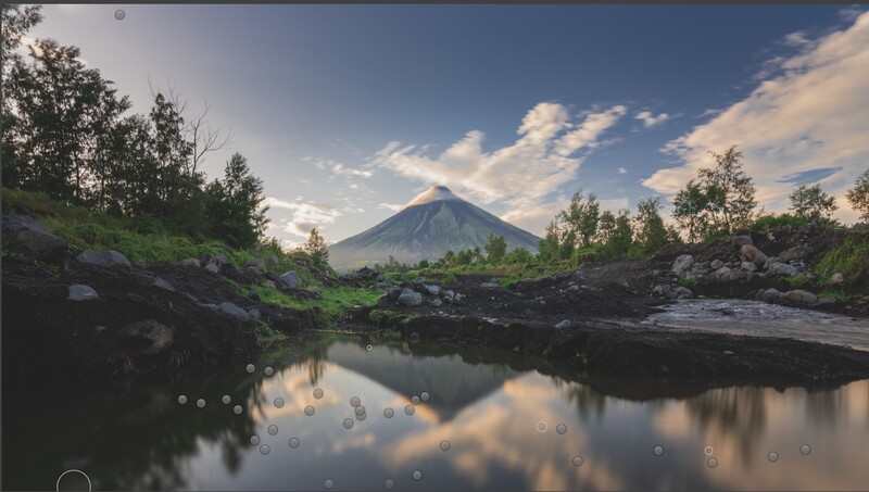 Conical volcano reflected in still water surrounded by lush green forest and rocky terrain.
