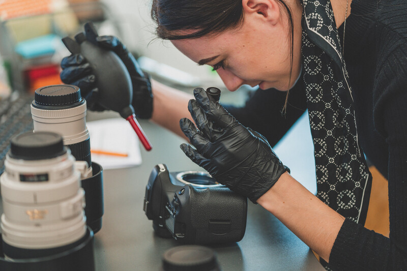 Female photographer wearing black gloves examines a camera lens at a workbench.