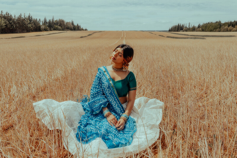 Woman in blue patterned dress sitting in harvested grain field with forest backdrop.