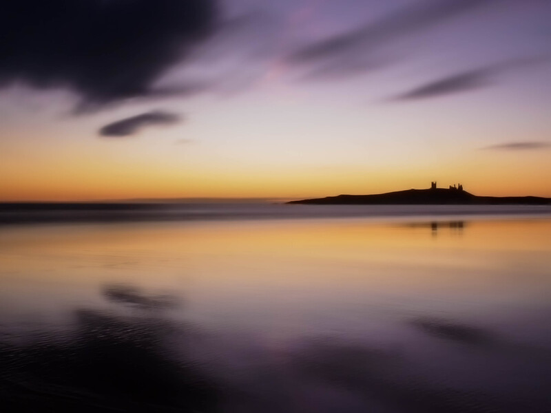 Coastal landscape at twilight with calm reflective water and distant structures silhouetted against purple and golden sky.