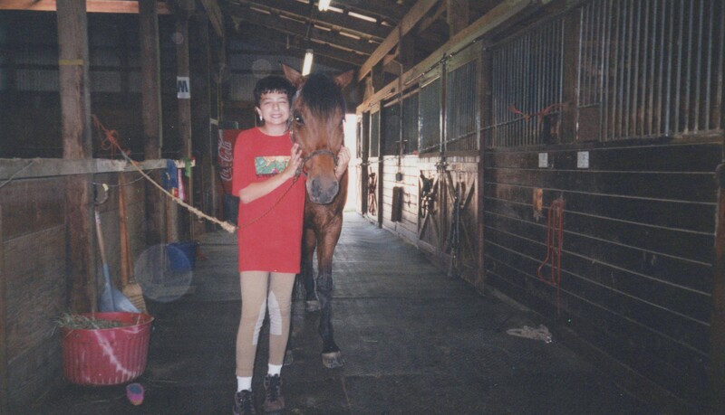 Young person in red shirt leading a brown horse down a stable corridor.