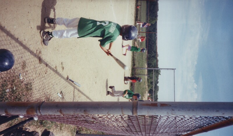 Young baseball player in green jersey at bat during youth game on dirt field.