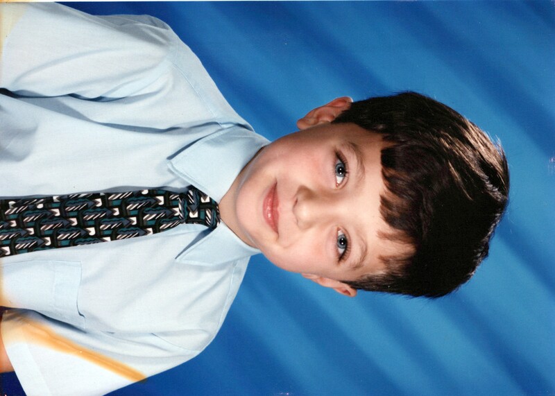 Young boy in light blue dress shirt and patterned tie against blue backdrop.