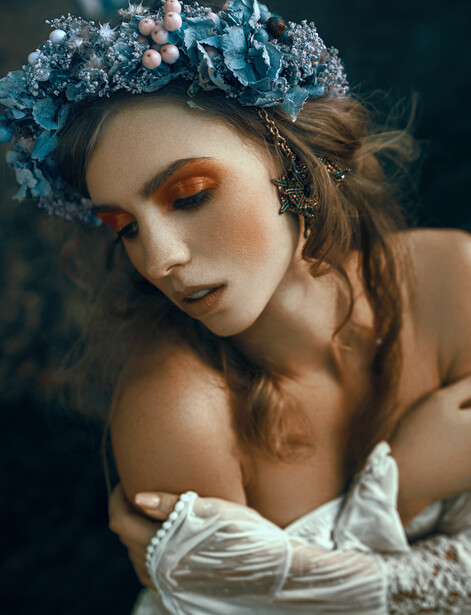 Woman with copper eyeshadow and textured blue floral headpiece, wearing white draped fabric.