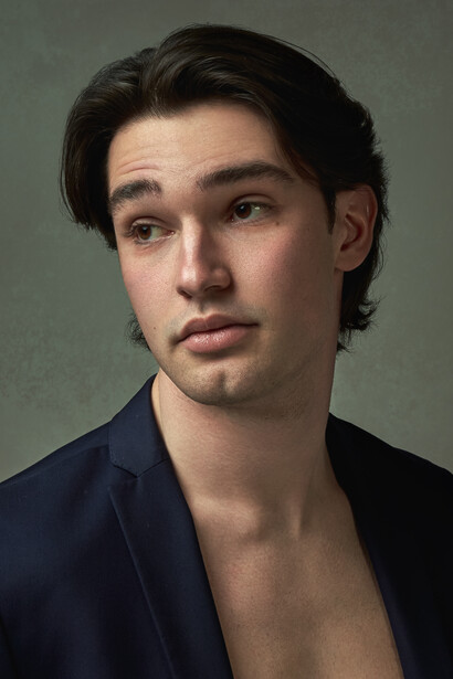 Young man with dark shoulder-length hair wearing an open navy blazer against a neutral gray background.