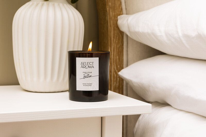 Lit black candle with white label on white shelf beside folded linens and ceramic vase.