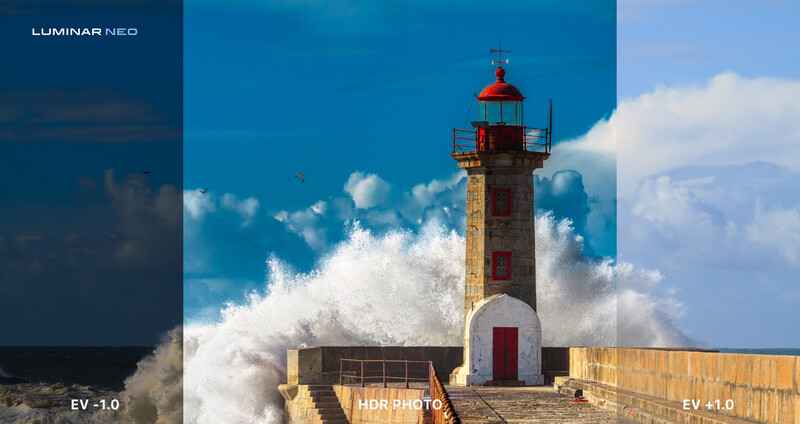 Lighthouse with dramatic ocean wave crashing against stone fortification under blue sky with white clouds.