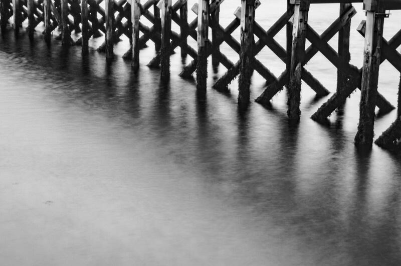 Wooden pier pilings extending into still water, captured in black and white with long exposure.
