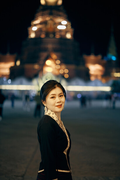 Woman in black blazer looking over shoulder with illuminated building in soft-focus background at night.