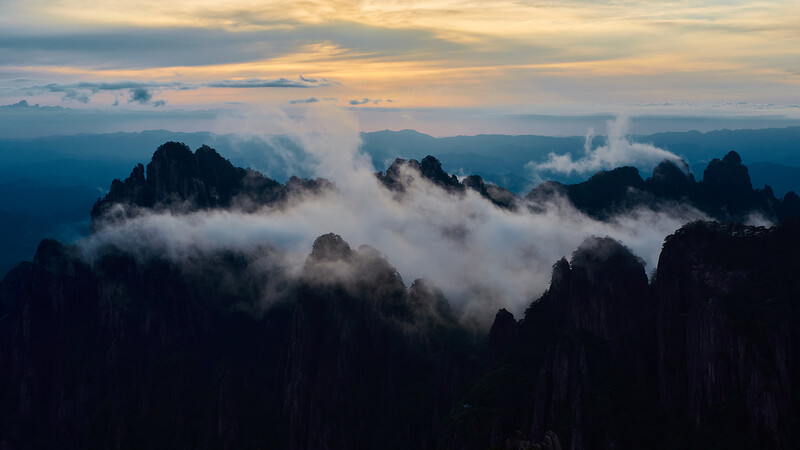 Dramatic mountain peaks shrouded in layered clouds at dawn, silhouetted against golden sky.