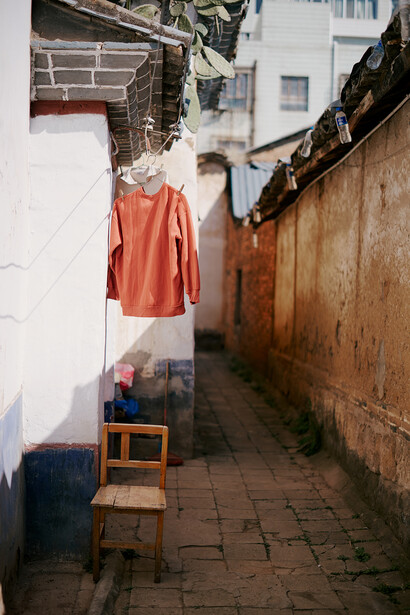 Orange shirt hanging on clothesline in narrow brick alley with wooden chair below.