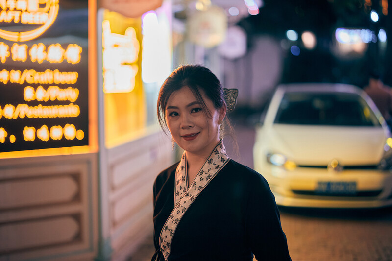 Woman in black blazer smiling at camera during evening street photography with warm storefront lighting.