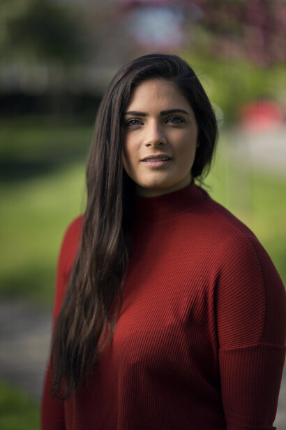 Woman with long dark hair wearing a burgundy sweater, photographed outdoors with a soft-focus green background.