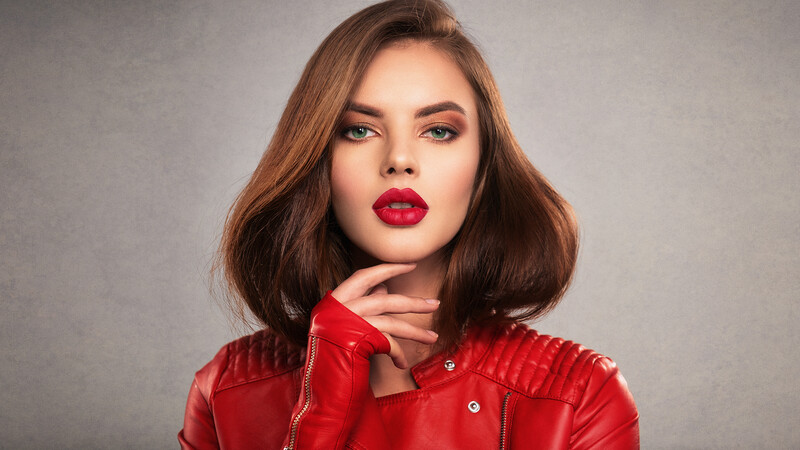 Woman with shoulder-length brown hair wearing red lipstick and a red quilted jacket against a neutral gray background.
