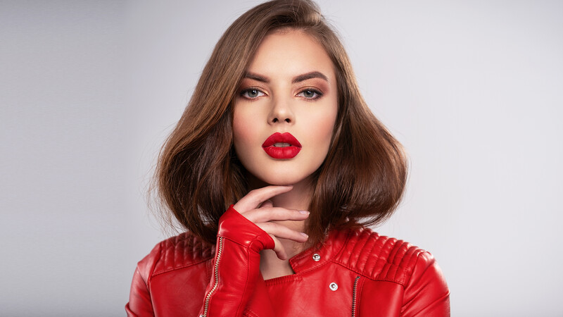 Woman with shoulder-length brown hair wearing bright red lipstick and a red quilted jacket against neutral background.