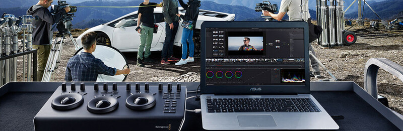Video editing workstation with control surface and laptop displaying color grading software, overlooking outdoor film production set with white vehicle and crew.