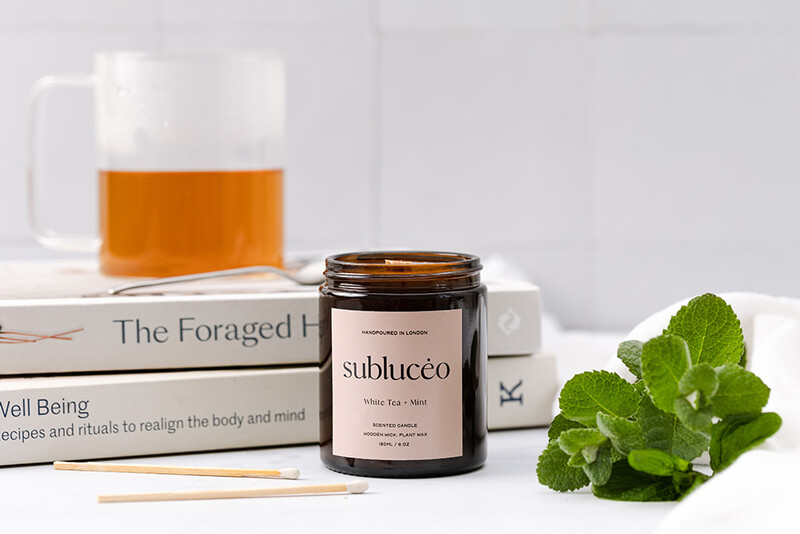 Flat lay composition with amber glass candle jar, stacked design books, fresh mint leaves, and mug of tea on white surface.