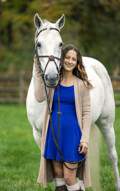 Young woman in blue dress and cardigan standing next to a white horse outdoors.