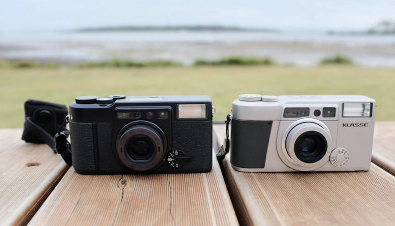 Two vintage film cameras displayed side-by-side on a wooden surface outdoors.