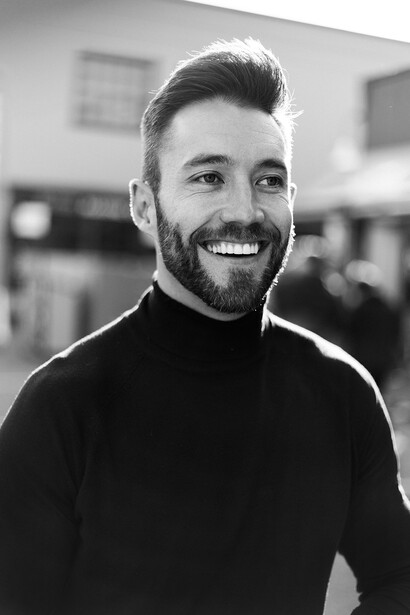 Man with dark hair and beard smiling at camera in black turtleneck against blurred indoor background.