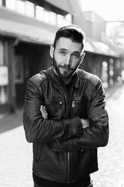 Man in black leather jacket with arms crossed, looking at camera on urban street.