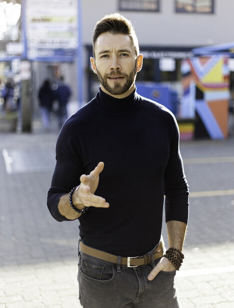Man in black turtleneck and gray pants gesturing toward camera in indoor public space.