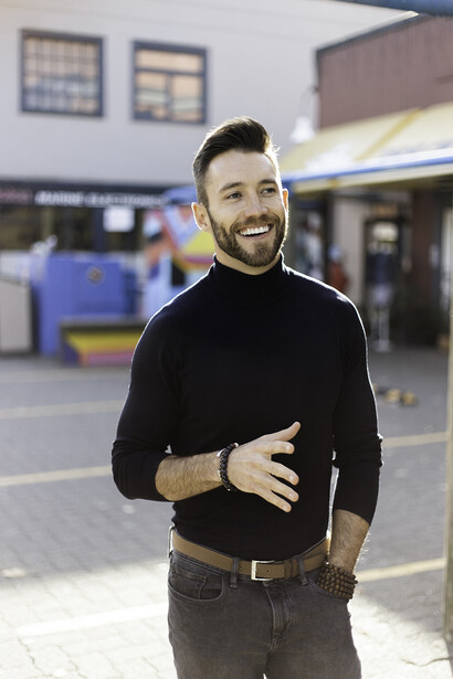 Man in black turtleneck and dark pants smiling outdoors in front of a storefront.