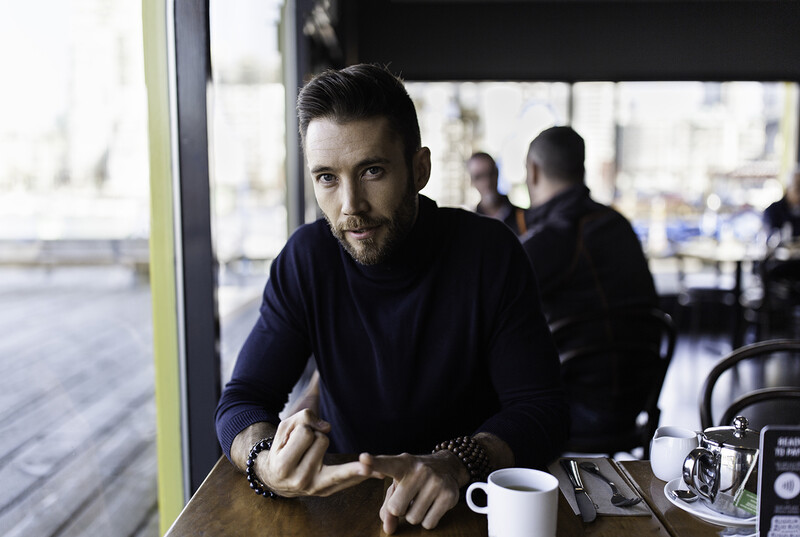 Man in dark navy hoodie seated at table near large window, looking directly at camera with serious expression.