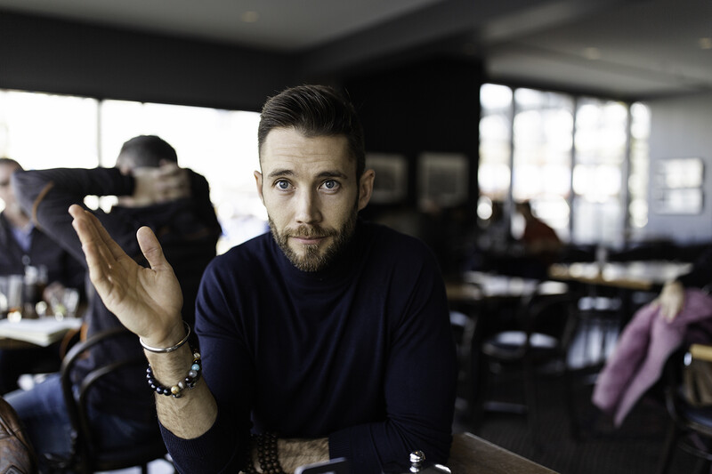 Bearded man in dark navy shirt gesturing while speaking in an office environment.