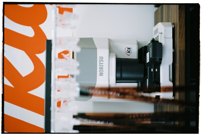 Noritsu photo lab printer displayed on wooden counter with orange branding signage overhead.