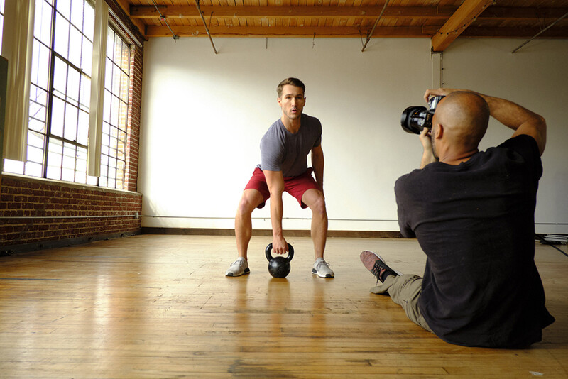 Photographer shooting fitness portrait of man holding kettlebell in industrial gym studio.