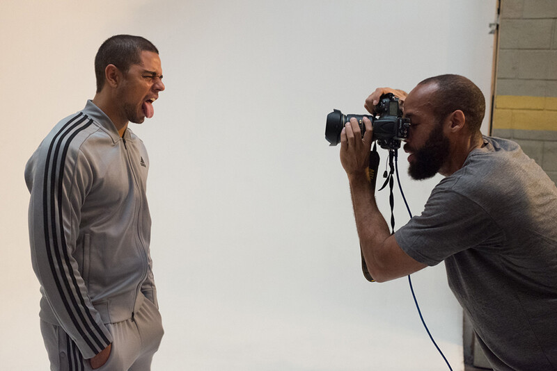 Photographer holding a DSLR camera with telephoto lens while capturing a portrait of a smiling man in athletic wear.