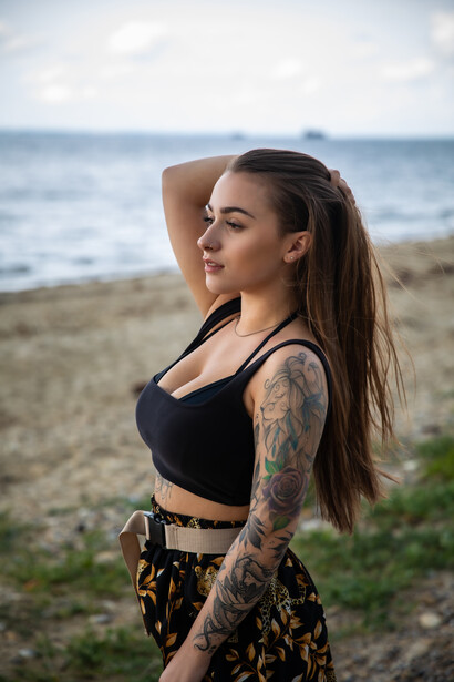 Woman with tattoos posing on a beach with the ocean and cloudy sky behind her.