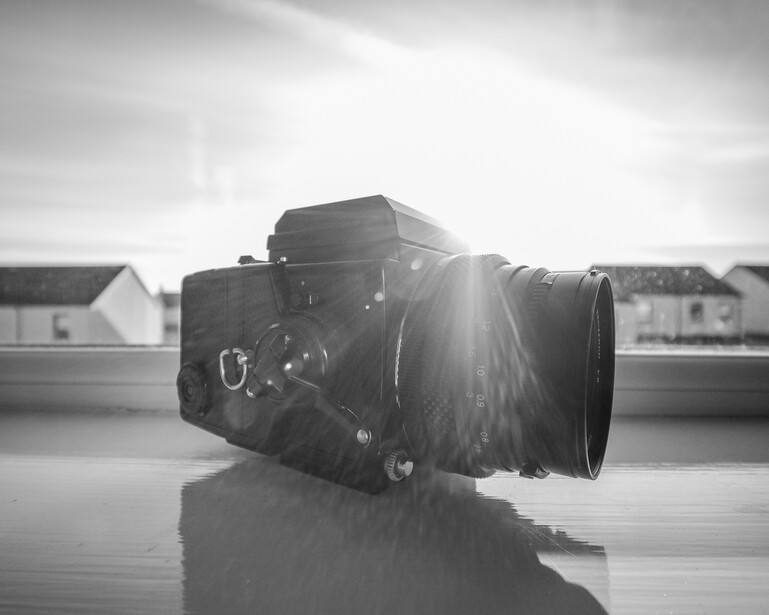 Medium format film camera with telephoto lens on reflective surface, backlit by bright sky.