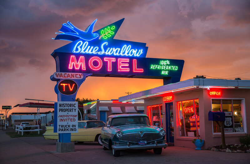 Vintage Blue Swallow Motel neon sign glowing at dusk with classic 1950s car parked in front.